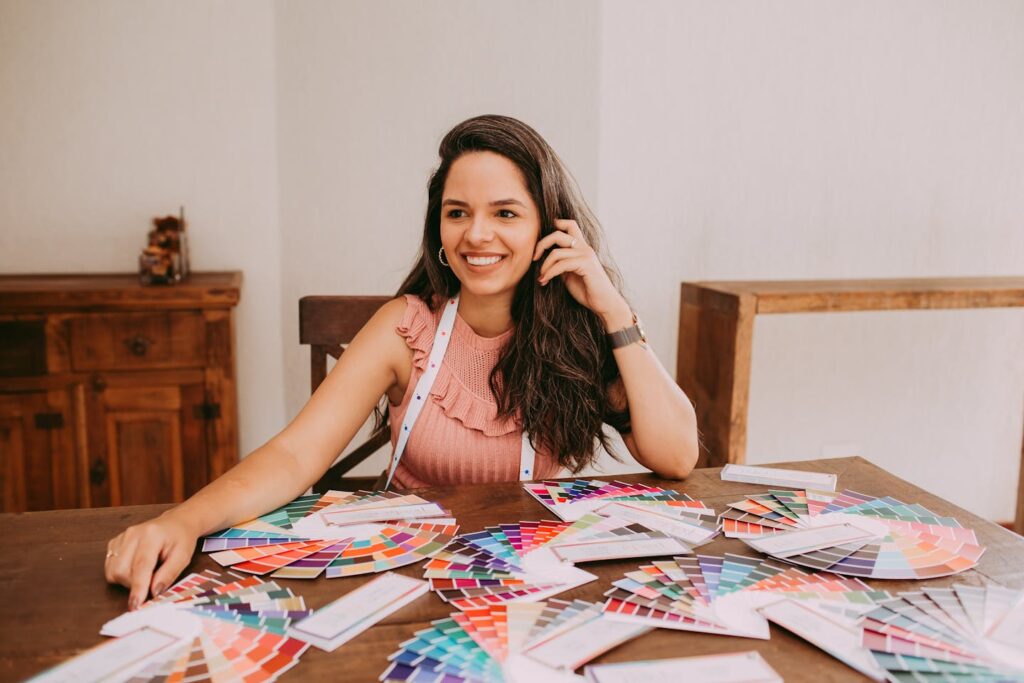 A woman with long hair, smiling while sitting at a table with various color swatches, suggesting creativity and design.